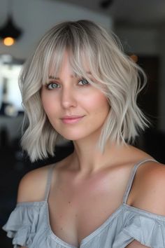 Woman with short, wavy blonde hair and bangs, wearing a gray, off-the-shoulder top, smiling softly. Angled Bobs With Bangs, Short Aline Bob, Bob With Side Swept Bangs, Short Aline, Angled Bob Haircut, Blonde Angled Bob, Bobs With Bangs, Grunge Haircut, Ash Blonde Bob