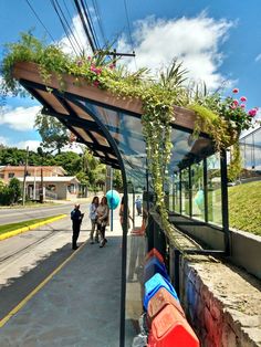 people are walking on the sidewalk next to a bus stop covered in plants and flowers