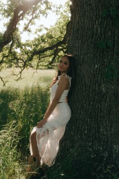 a woman in a white dress leaning against a tree