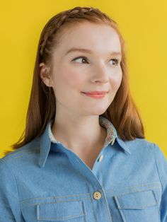 a woman with red hair wearing a denim shirt and smiling at the camera, against a yellow background