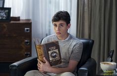 a young man sitting at a desk holding a book in front of him and looking surprised