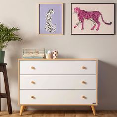 a white dresser with two pictures on the wall above it and a potted plant next to it