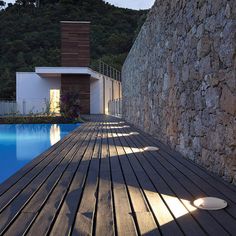 a wooden deck next to a swimming pool in front of a stone wall and house