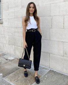 a woman in black pants and white top holding a handbag while standing next to a wall