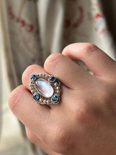 “Art Deco” was an era marked by extravagance and innovation. The Art Deco style itself was an eclectic mix of heavy geometric shapes, with curved ornamental elements. The resulting silhouettes were often monolithic but elegant, with applied decorative elements. This Moonstone and Sapphire Edwardian Conversion Ring is a wonderful example of this iconic Art Deco style. Crafted in 20 carat white gold, this ring immediately captures the gaze with its dazzling array of miniature pearls and sapphire stones haloing a truly luminescent cabochon moonstone. What makes this treasure particularly special is that it is a conversion piece; the beautiful moonstone center was once part of an antique brooch. Now, with the use of a 14 carat gold replica shank, it has been transformed into the glorious ring Luxury Silver Moonstone Ring, Formal Fusion Rings With Accent Stones, Formal Fusion Style Rings With Accent Stones, Modernist Gemstone Ring Jewelry, Art Deco Multi-stone Oval Jewelry, Elegant Formal Multi-stone Moonstone Ring, Unique Oval Moonstone Ring For Formal Occasions, Luxury Oval Hand Forged Jewelry, Luxury Silver Oval Moonstone Ring