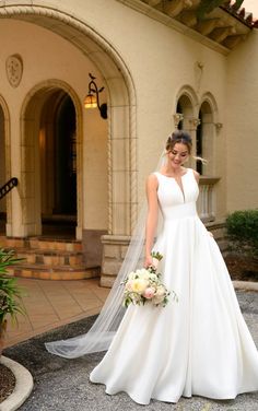 a woman standing in front of a building wearing a wedding dress and holding a bouquet