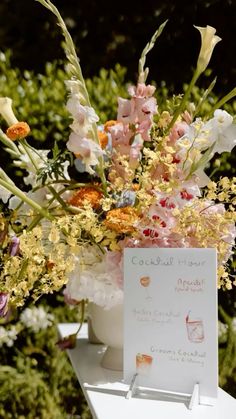 an arrangement of flowers in a vase with a sign on the table next to it