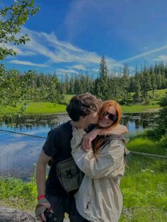 two people standing next to each other in front of a lake with trees and grass