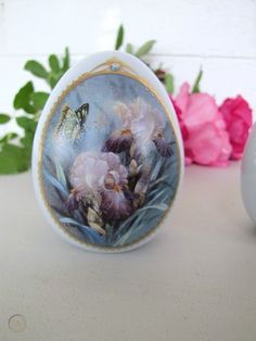 an egg with flowers painted on it sitting next to a vase and pink roses in the background