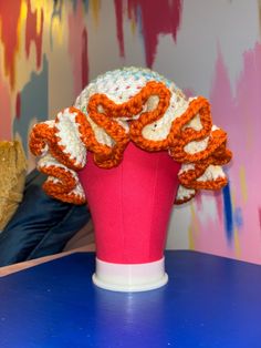 an orange and white knitted hat sitting on top of a pink vase in front of a colorful wall