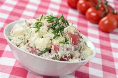 a white bowl filled with potato salad on top of a checkered table cloth next to tomatoes