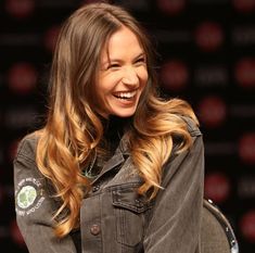 a woman with long hair smiling and wearing a denim jacket over her shoulders in front of a black background