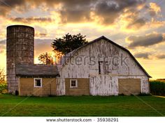 an old barn and silo with the sun setting in the sky behind it on a farm