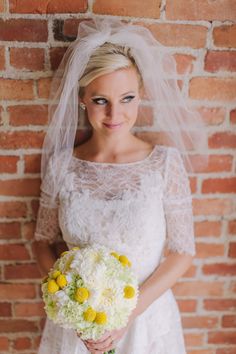 a woman in a wedding dress standing next to a brick wall holding a bouquet of flowers