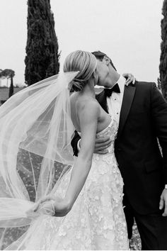 a bride and groom kissing in front of some trees with their veil blowing in the wind