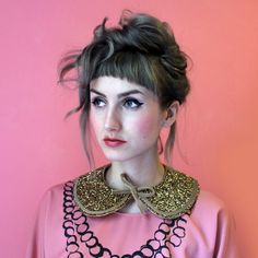 a woman wearing a pink top with gold collar and beads on her neck, standing against a pink wall