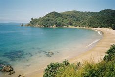 a sandy beach with clear blue water surrounded by green hills and trees in the background