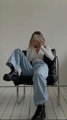 a woman sitting in a chair taking a selfie with her cell phone while wearing jeans and boots