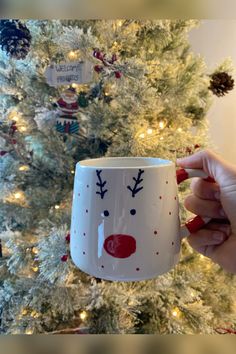 someone holding up a coffee mug with reindeer designs on it in front of a christmas tree