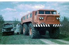 an orange monster truck driving down a dirt road next to a black car on the other side