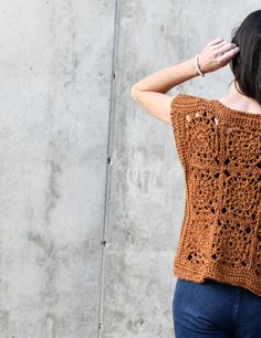 a woman wearing a brown crochet top and blue jeans stands in front of a concrete wall