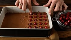 two hands reaching for chocolate in a pan with cherries on it and another hand touching the batter