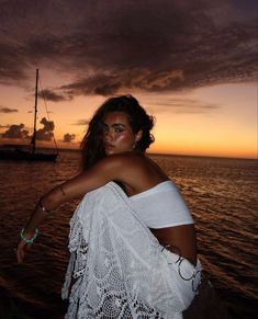 a woman in white dress sitting on the edge of water at sunset with ship in background