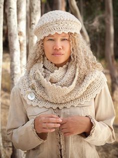 a woman wearing a hat and scarf standing in the woods with her hands folded over her chest