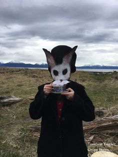 a person wearing a cat mask and holding a bowl in front of their face while standing on the grass