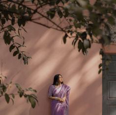 a woman in a purple sari leaning against a pink wall with her hands on her hips