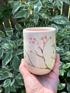 a hand holding a white cup with pink flowers on it in front of some bushes