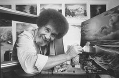 a man with an afro is smiling in front of some paintings and holding a paintbrush