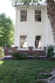 a white house with two windows and a tree in the front yard on a sunny day