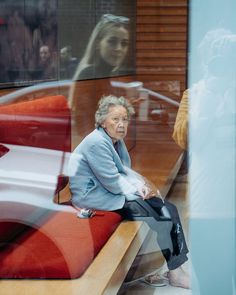 an older woman sitting on a bench in front of a glass window looking at her reflection