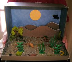 a child's hand holding a cardboard box with desert scene inside