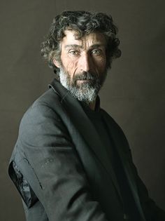 a man with curly hair and beard in a suit poses for the camera while wearing a black shirt