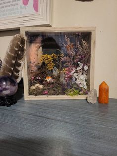 there is a glass frame with flowers and plants in it next to a candle on the table