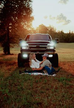 a man and woman sitting in the grass next to a pickup truck with headlights on