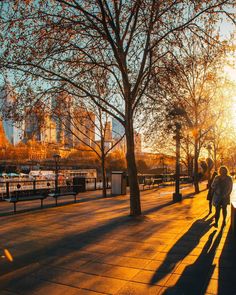 two people walking down the sidewalk in front of some trees and buildings at sunset or sunrise