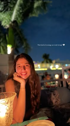 a woman sitting at a table with a lit candle in front of her and palm trees behind her