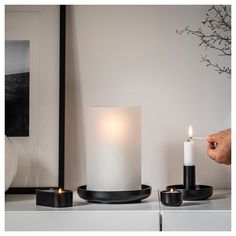 a person lighting a candle on top of a white shelf next to two black candles