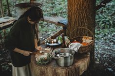 a woman preparing food on top of a wooden table next to a tree in the woods