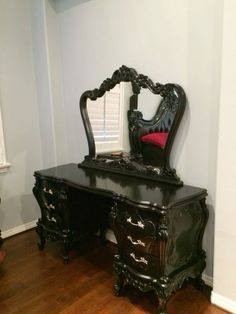 an ornate black vanity with mirror and red cushion on the floor in front of a white wall