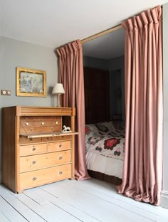 a bed sitting next to a wooden dresser in a bedroom under a window with pink drapes