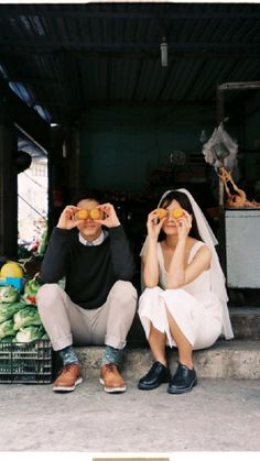 two people sitting on the ground with fruit in front of their eyes and one person holding an orange up to his face