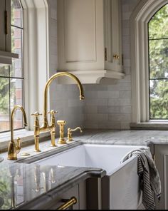 a kitchen sink with gold faucets and marble counter tops in front of two windows