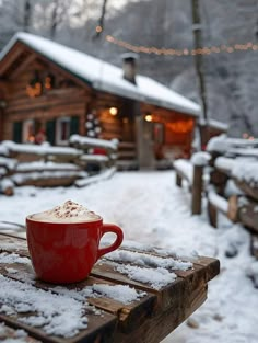 a red coffee cup sitting on top of a wooden table covered in snow next to a log cabin