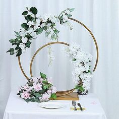 a white table topped with flowers and a gold circular frame filled with greenery on top of a white cloth covered table