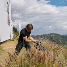 a man standing on top of a lush green hillside next to tall grass and a building