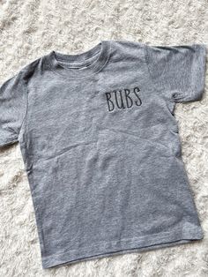 This sweet and simple brother shirt is embroidered in charcoal grey thread with the sweet name "bubs" on the left chest. This shirt is perfect for any little boy whether they are already a brother, about to become one or goes by the name "bubs".  This cozy sibling t-shirt can be paired with one of our baby announcement signs to provide the perfect announcement that you are adding to your family! Details: Thread is polyester Embroidery Wash and Care: -Always wash your garment before use -Wash ins Casual Gray Tops With Embroidered Text, Casual Gray Top With Embroidered Text, Basic Gray Shirt With Letter Print, Gray Cotton Tops With Name Print, Gray Short Sleeve Top With Funny Text, Gray Embroidered Text Crew Neck Top, Big Brother T Shirt, Big Brother Announcement Tshirt, Big Bro Little Bro Shirts